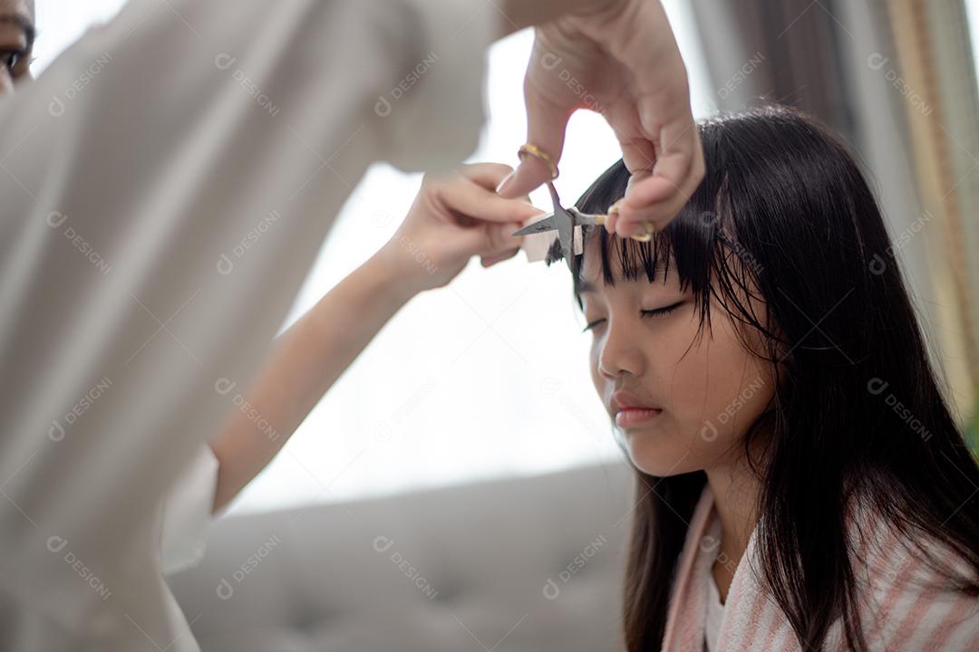 Mãe asiática cortando cabelo para a filha na sala de estar em casa enquanto fica em casa segura do Covid-19 Coronavirus durante o bloqueio. Conceito de auto-quarentena e distanciamento social.