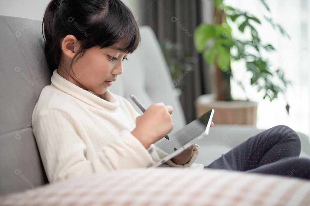 Menina usando tablet jogando na internet, criança sentada no sofá assistindo ou conversando com um amigo online, criança relaxando na sala de manhã, crianças com conceito de nova tecnologia
