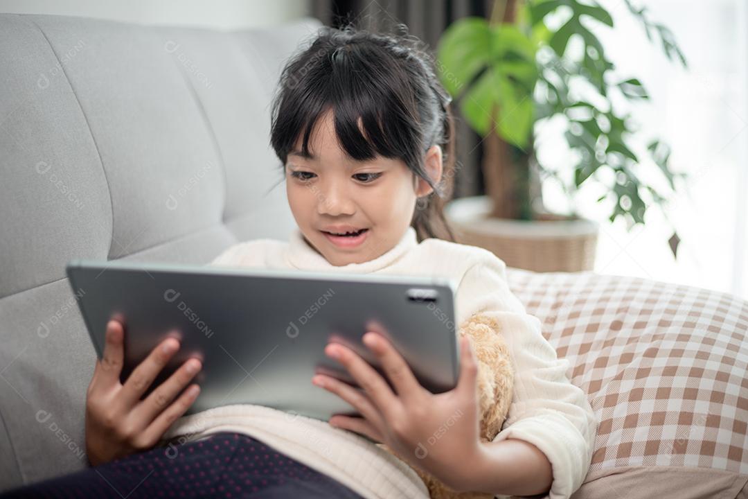 Menina usando tablet jogando na internet, criança sentada no sofá assistindo ou conversando com um amigo online, criança relaxando na sala de manhã, crianças com conceito de nova tecnologia