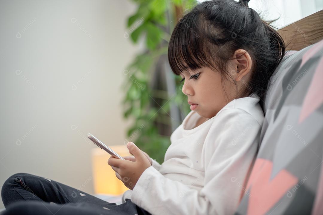 Garotinha bonitinha assistindo vídeo no smartphone com carinha sozinha na cama, criança usando celular com carinha feliz em casa. Fique em casa em quarentena coronavírus COVID-19 prevenção pandêmica.