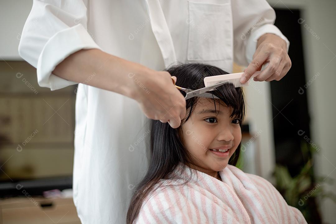 Mãe asiática cortando cabelo para a filha na sala de estar em casa enquanto fica em casa segura do Covid-19 Coronavirus durante o bloqueio. Conceito de auto-quarentena e distanciamento social.