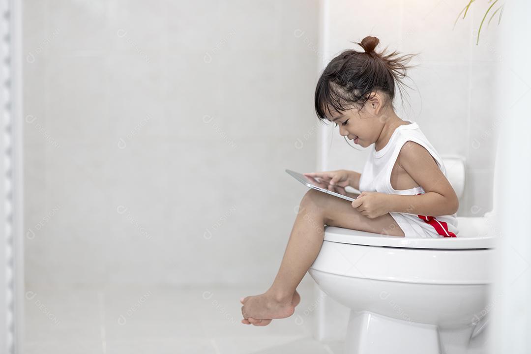 criança sentada no vaso sanitário segurando o tablet. conceito de smartphone viciado em criança