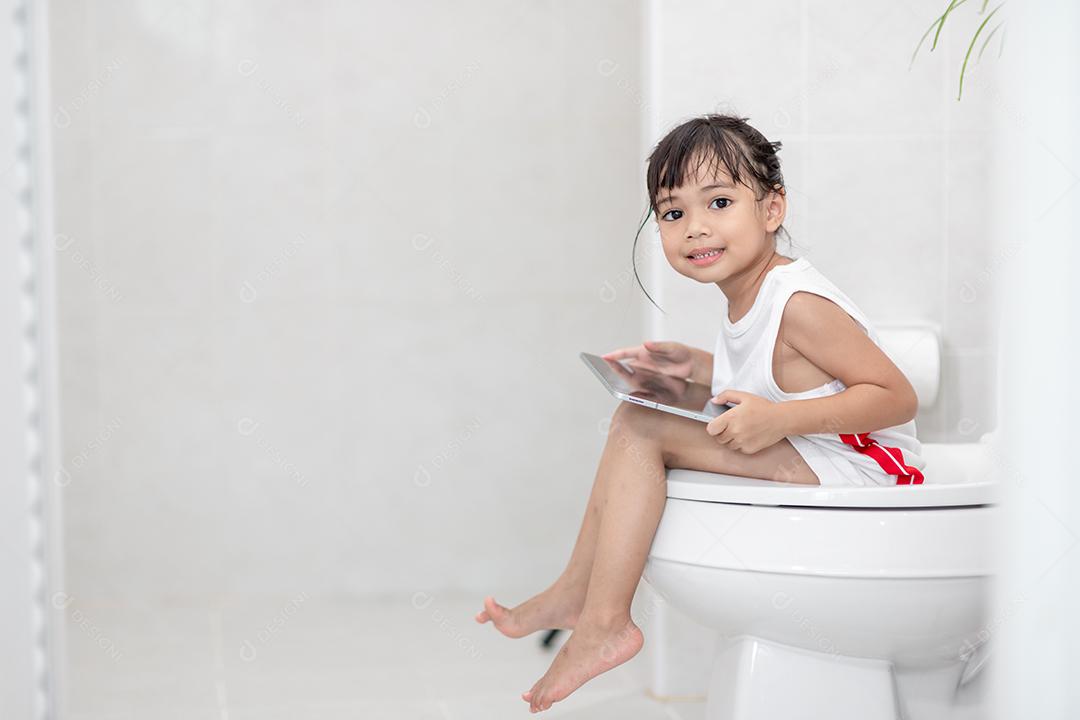 criança sentada no vaso sanitário segurando o tablet. conceito de smartphone viciado em criança