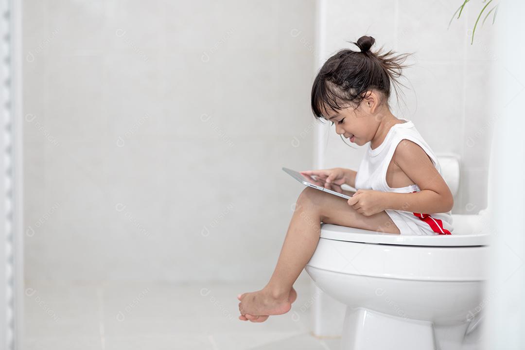 criança sentada no vaso sanitário segurando o tablet. conceito de smartphone viciado em criança