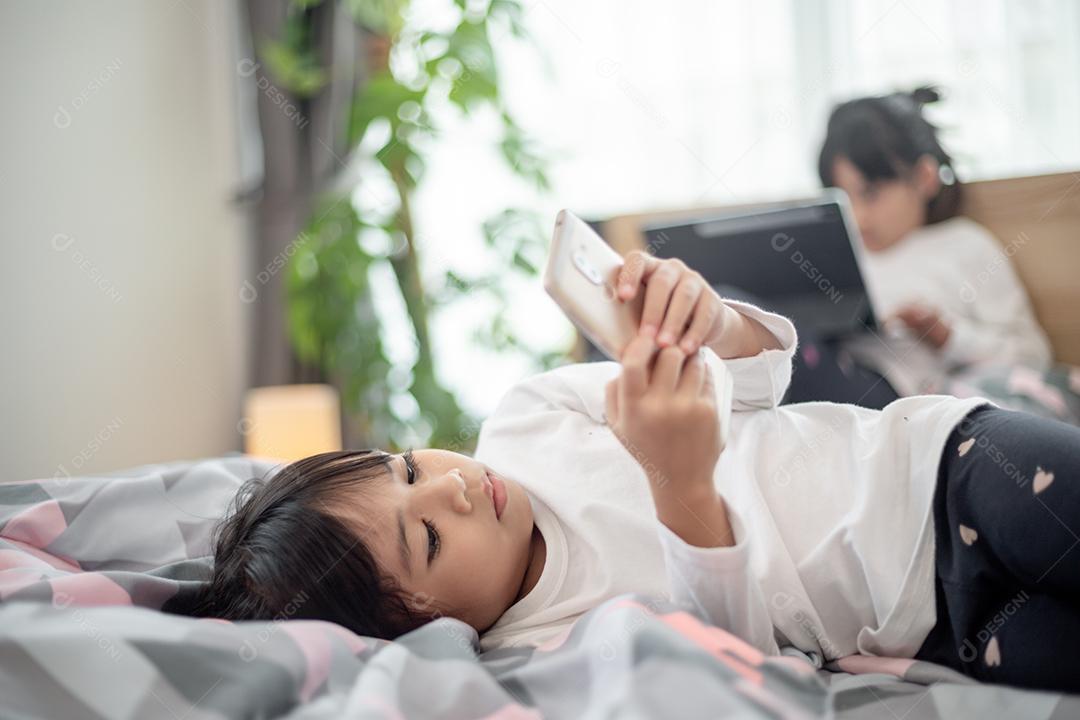 Garotinha bonitinha assistindo vídeo no smartphone com carinha sozinha na cama, criança usando celular com carinha feliz em casa. Fique em casa em quarentena coronavírus COVID-19 prevenção pandêmica.