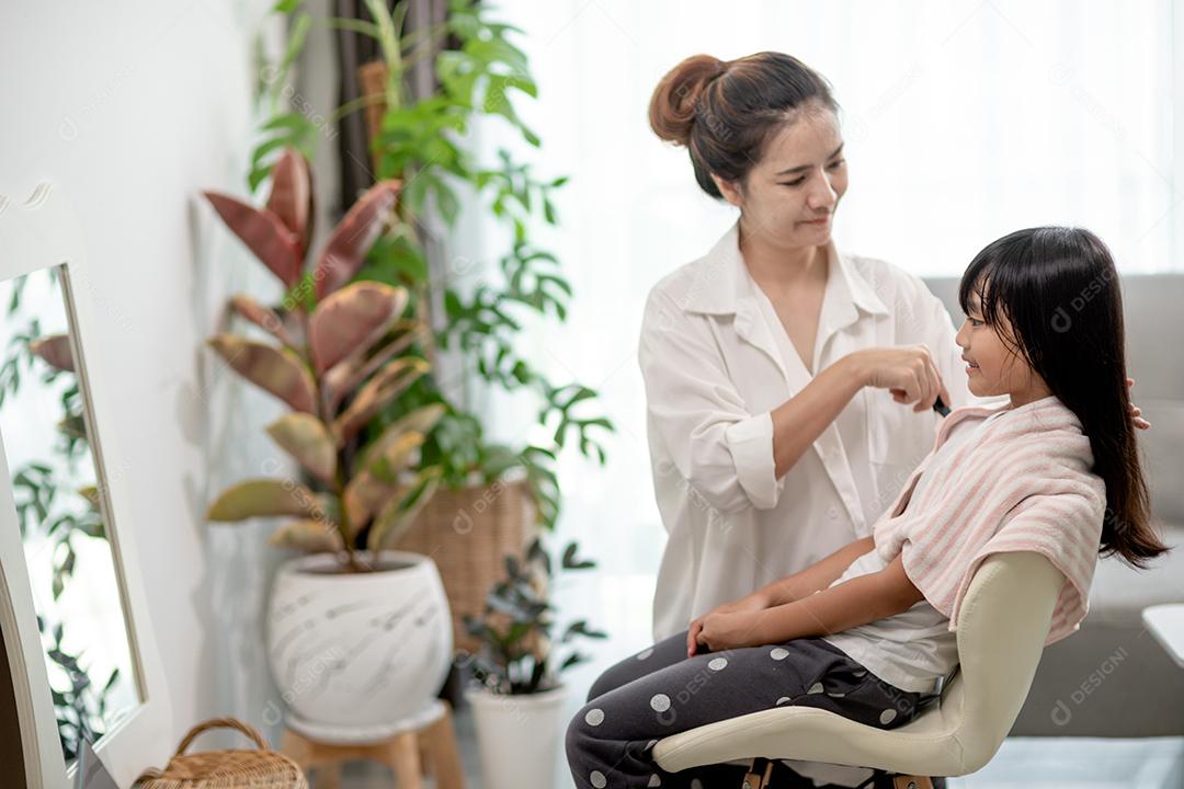 Mãe asiática cortando cabelo para a filha na sala de estar em casa enquanto fica em casa segura do Covid-19 Coronavirus durante o bloqueio. Conceito de auto-quarentena e distanciamento social.
