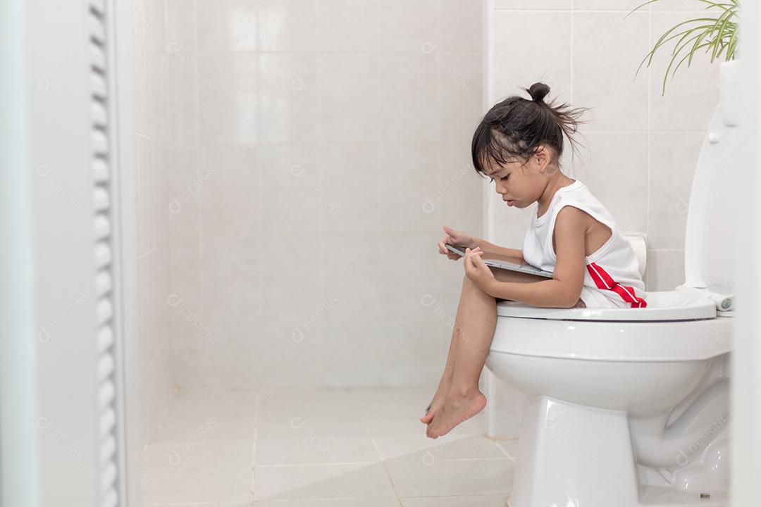 criança sentada no vaso sanitário segurando o tablet. conceito de smartphone viciado em criança