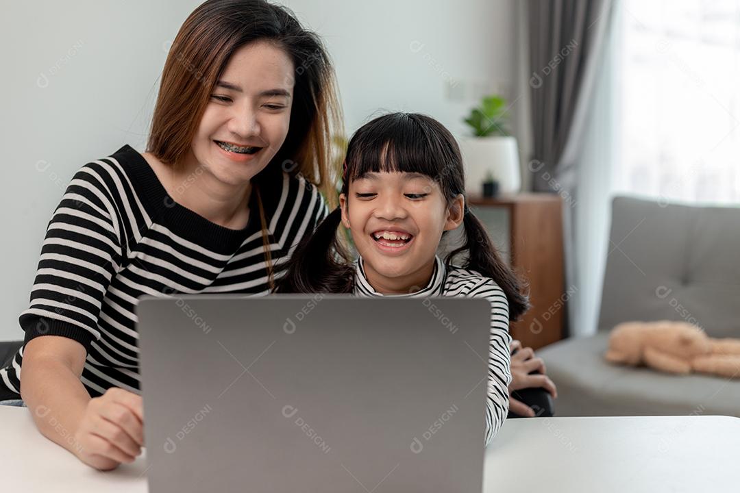 Garotinha asiática aprendendo aula online em casa com a mãe. A criança pré-escolar usa o laptop para fazer a lição de casa, o ensino em casa do professor da escola pela internet remota digital com o apoio da mãe.