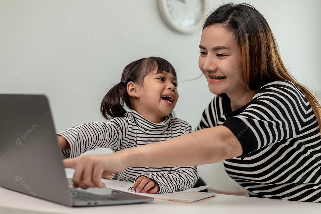 Garotinha asiática aprendendo aula online em casa com a mãe. A criança pré-escolar usa o laptop para fazer a lição de casa, o ensino em casa do professor da escola pela internet remota digital com o apoio da mãe.