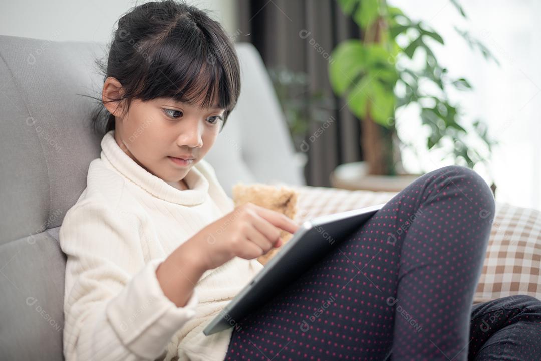 Menina usando tablet jogando na internet, criança sentada no sofá assistindo ou conversando com um amigo online, criança relaxando na sala de manhã, crianças com conceito de nova tecnologia