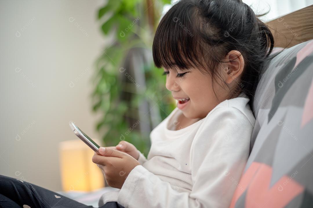 Garotinha bonitinha assistindo vídeo no smartphone com carinha sozinha na cama, criança usando celular com carinha feliz em casa. Fique em casa em quarentena coronavírus COVID-19 prevenção pandêmica.