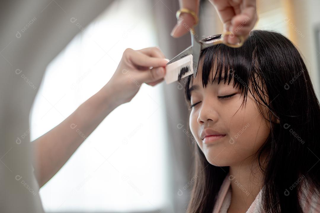 Mãe asiática cortando cabelo para a filha na sala de estar em casa enquanto fica em casa segura do Covid-19 Coronavirus durante o bloqueio. Conceito de auto-quarentena e distanciamento social.