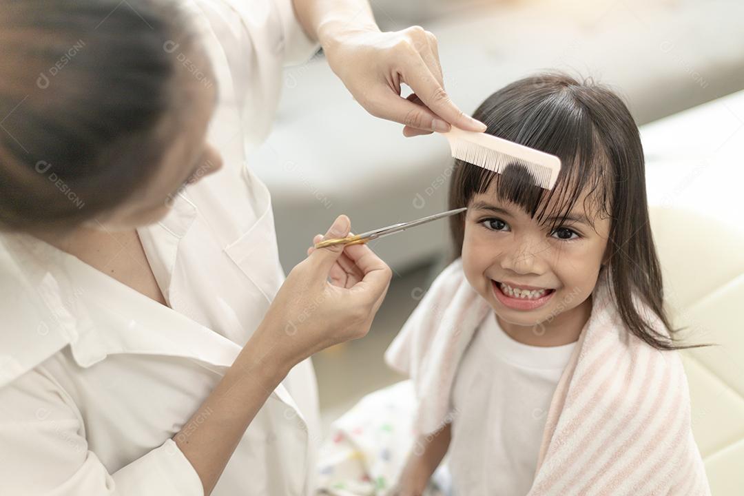 Mãe asiática cortando cabelo para a filha na sala de estar em casa enquanto fica em casa segura do Covid-19 Coronavirus durante o bloqueio. Conceito de auto-quarentena e distanciamento social.