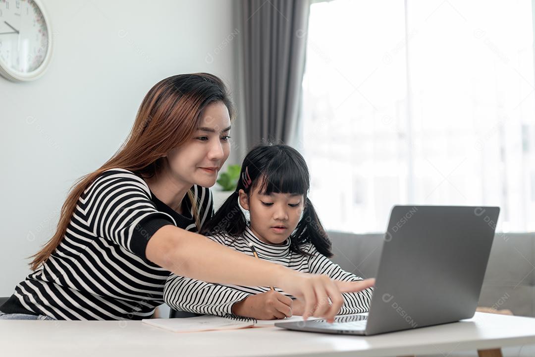 Garotinha asiática aprendendo aula online em casa com a mãe. A criança pré-escolar usa o laptop para fazer a lição de casa, o ensino em casa do professor da escola pela internet remota digital com o apoio da mãe.