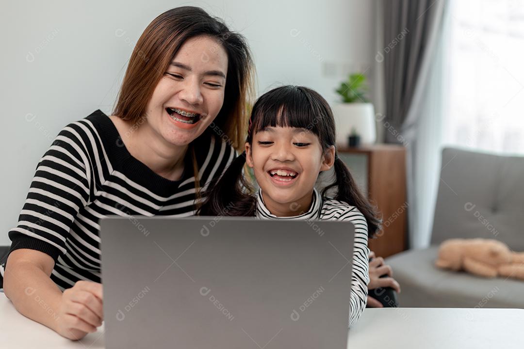 Garotinha asiática aprendendo aula online em casa com a mãe. A criança pré-escolar usa o laptop para fazer a lição de casa, o ensino em casa do professor da escola pela internet remota digital com o apoio da mãe.