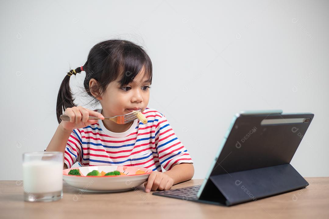 Menina adorável criança da Ásia almoçando enquanto assiste a um filme do tablet. Uma criança asiática comendo o jantar e os olhos estão olhando desenhos animados do Tablet. Semana Nacional de Conscientização sobre Transtornos Alimentares.
