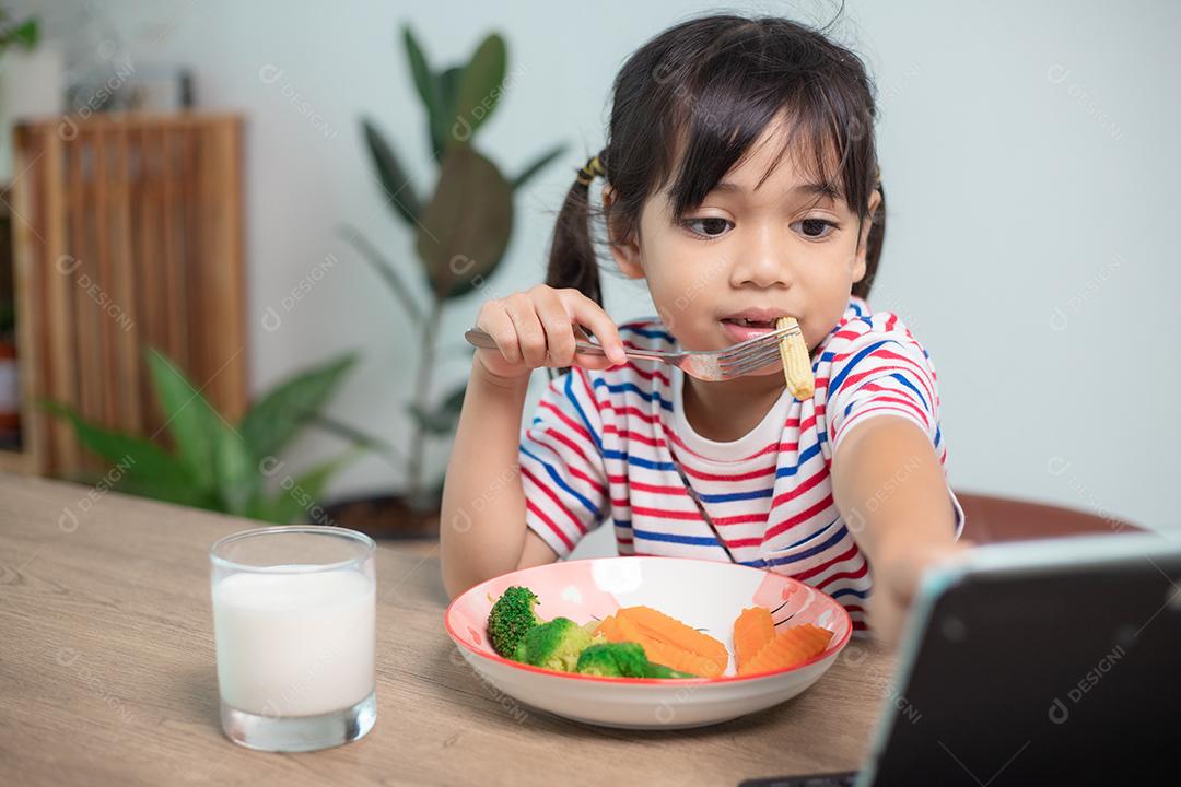Menina adorável criança da Ásia almoçando enquanto assiste a um filme do tablet. Uma criança asiática comendo o jantar e os olhos estão olhando desenhos animados do Tablet. Semana Nacional de Conscientização sobre Transtornos Alimentares.