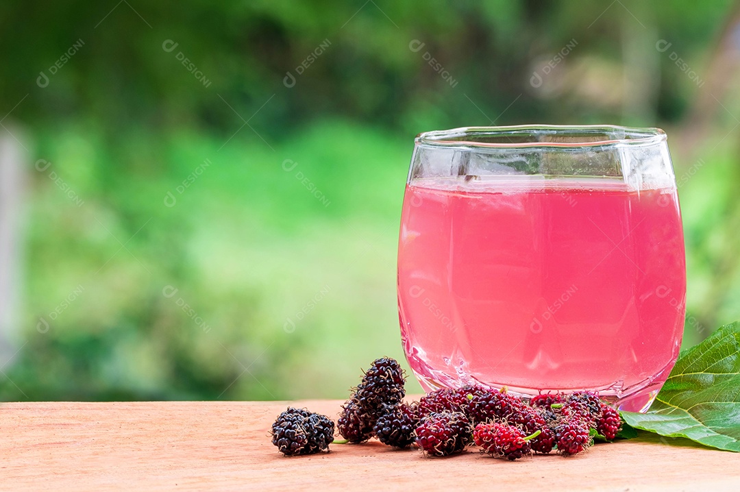 suco de amora no copo e fruta de amora na folha de amoreira colocada na placa de madeira contra o fundo verde da planta.
