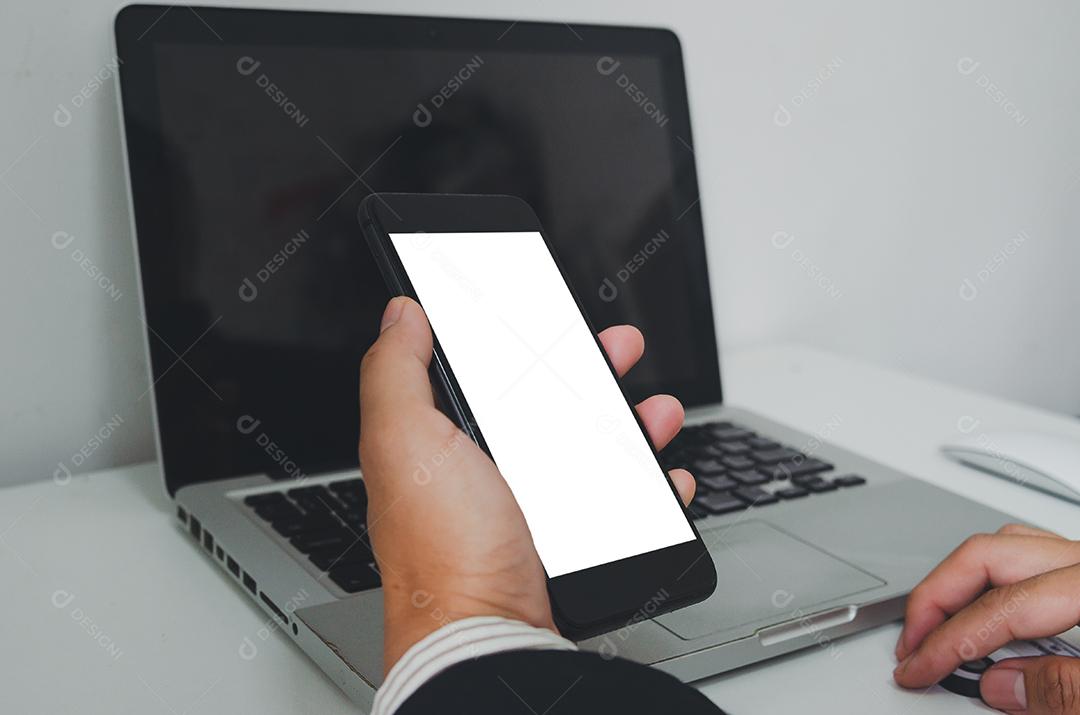 Empresário segurando telefone inteligente simulado tela branca e computador portátil na mesa.