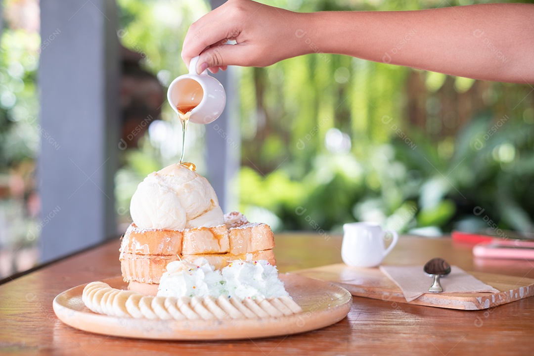 Mão de uma mulher segurando uma xícara de mel derramando sobre torradas de mel em uma bandeja de madeira, sobre uma mesa de madeira.