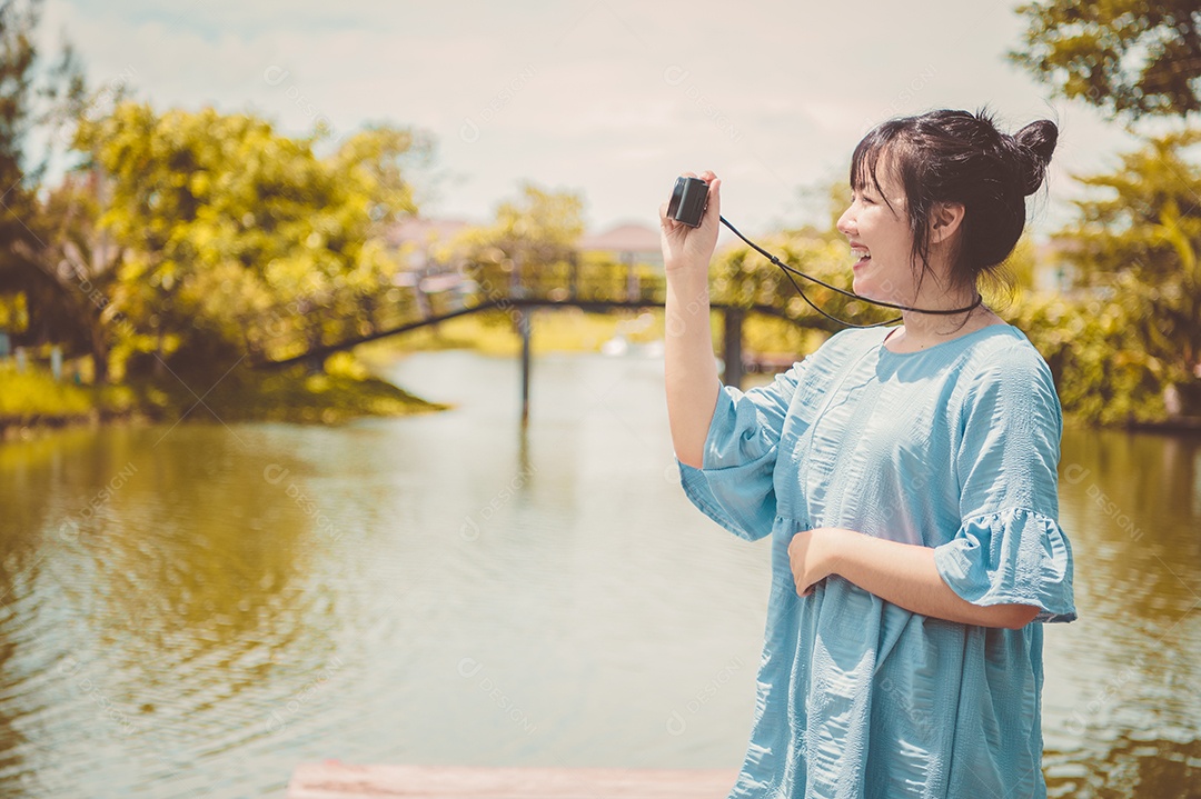 Mulher asiática de vestido azul em parque público carregando câmera digital sem espelho e tirando foto sem máscara facial de bom humor. Estilo de vida de pessoas e conceito de lazer. Viagens ao ar livre e tema da natureza.
