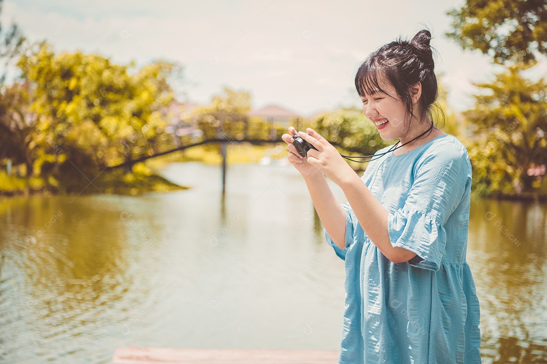 Mulher asiática de vestido azul em parque público carregando câmera digital sem espelho e tirando foto sem máscara facial de bom humor. Estilo de vida de pessoas e conceito de lazer. Viagens ao ar livre e tema da natureza.