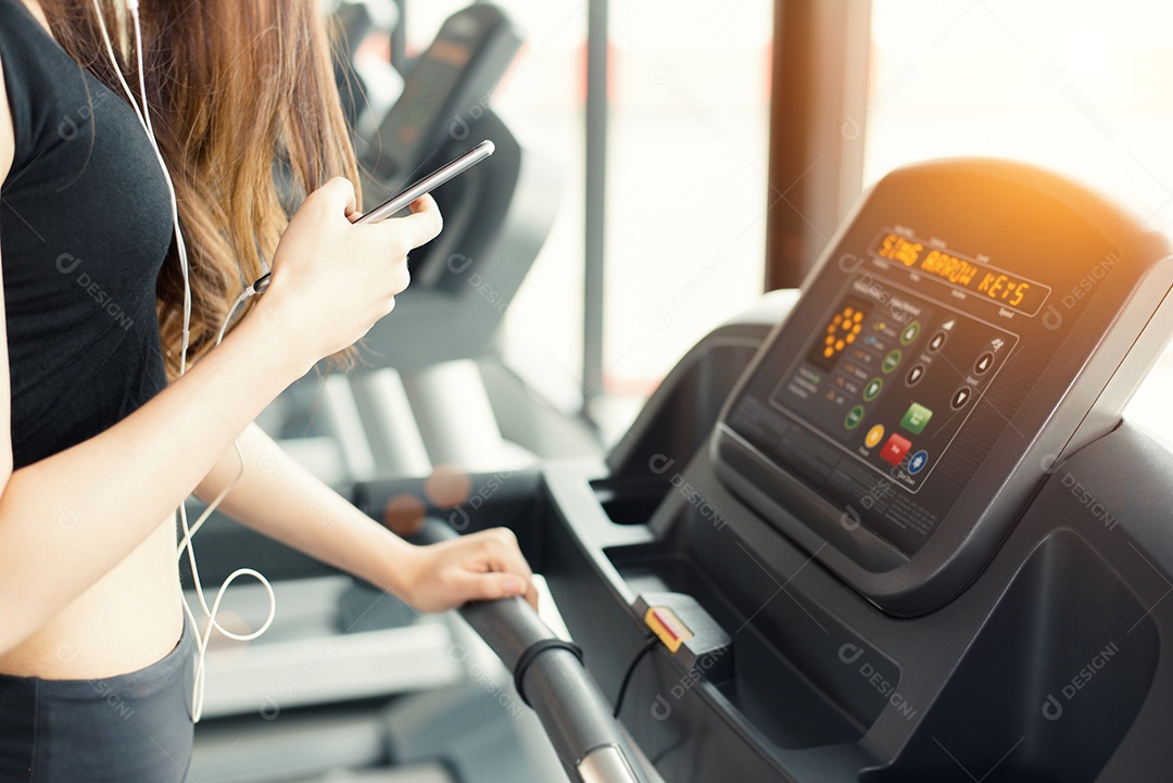 Mulher asiática usando telefone inteligente durante treino ou treinamento de força na academia de ginástica na esteira. Relaxe e o conceito de tecnologia. Exercício de esportes e tema de cuidados de saúde. Humor feliz e confortável.