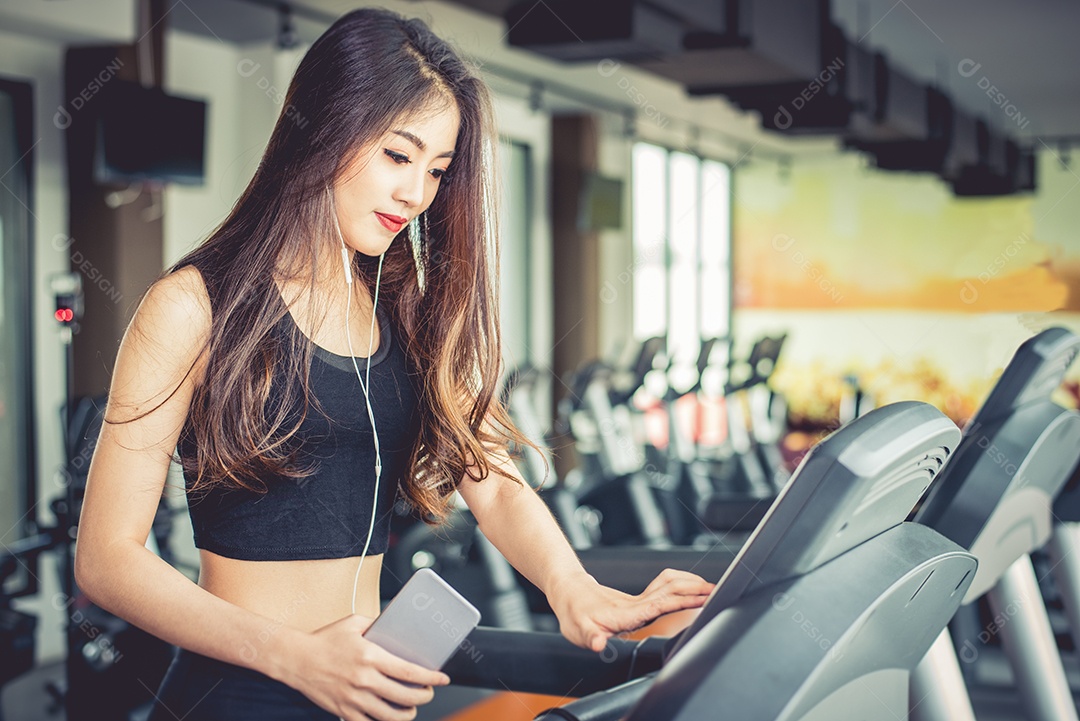 Mulher asiática usando telefone inteligente durante treino ou treinamento de força na academia de ginástica na esteira. Relaxe e o conceito de tecnologia. Exercício de esportes e tema de cuidados de saúde. Humor feliz e confortável.