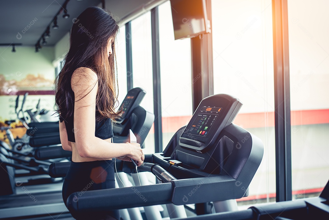 Mulher asiática usando telefone inteligente durante treino ou treinamento de força na academia de ginástica na esteira. Relaxe e o conceito de tecnologia. Exercício de esportes e tema de cuidados de saúde. Humor feliz e confortável.