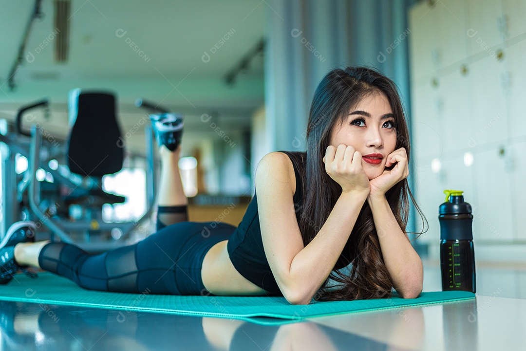 Mulher esportiva deitada e relaxando no tapete de ioga na academia de ginástica com fundo de equipamentos esportivos. Conceito de exercício de treinamento de beleza e treino. Construção corporal e tema Força.