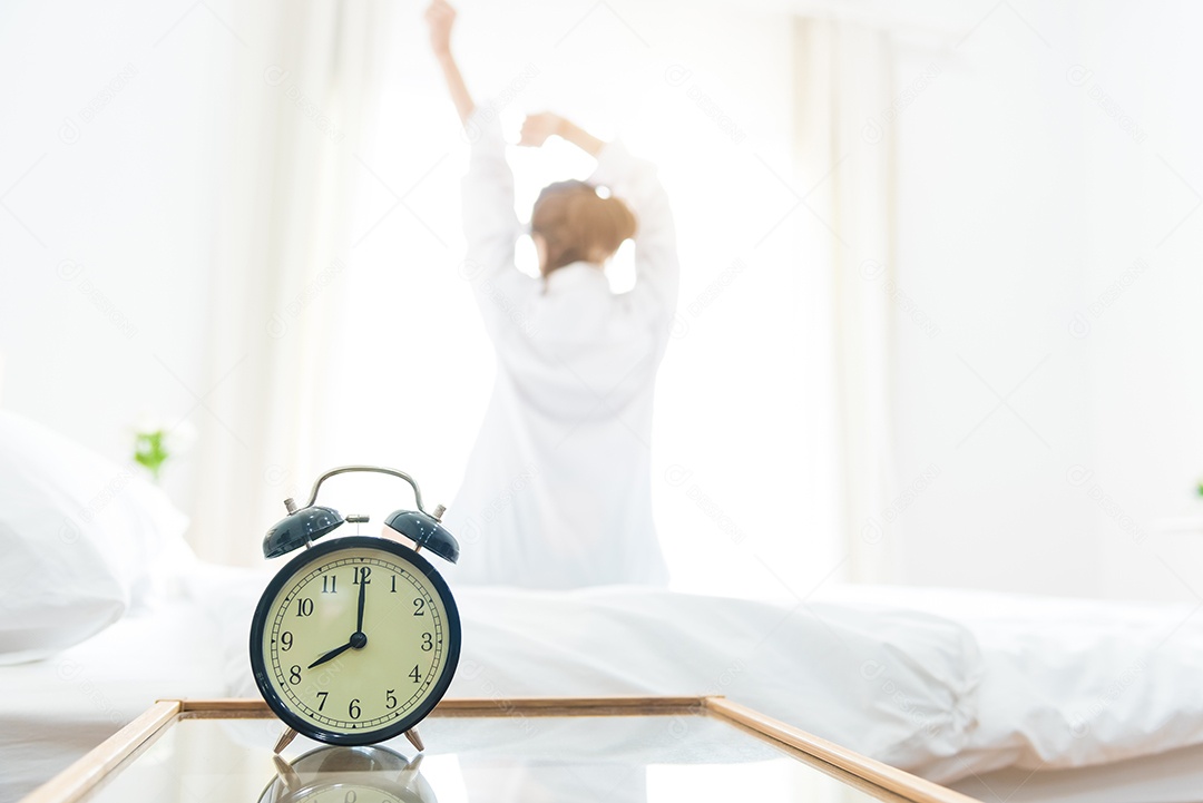 Vista traseira da mulher estendendo-se pela manhã depois de acordar na cama perto da janela com despertador.