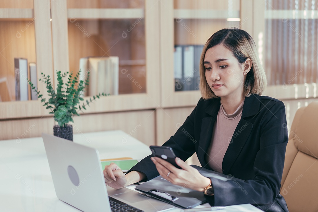 Gerente feminina asiática assistindo telefone celular e trabalhando com laptop