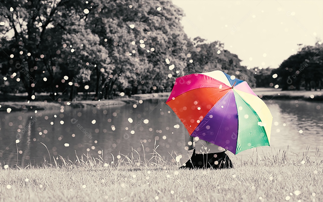 Guarda-chuva de arco-íris colorido segurando por mulher sentada no campo de grama perto do rio ao ar livre com cheio de natureza e chuva.