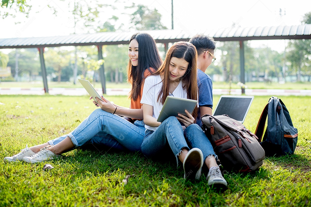 Grupo de estudantes universitários asiáticos usando tablet e laptop no campo de grama ao ar livre.