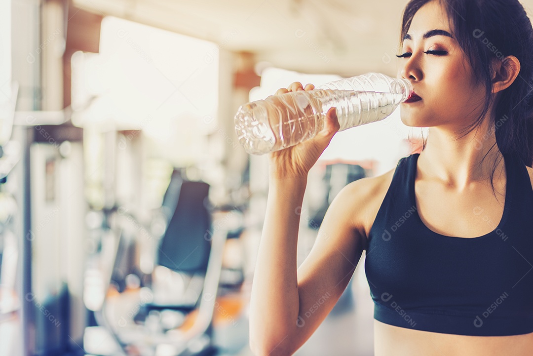 Mulher asiática bebendo água potável pura para frescura após treino ou treinamento de exercícios na academia de ginástica com fundo de equipamentos de ginástica.