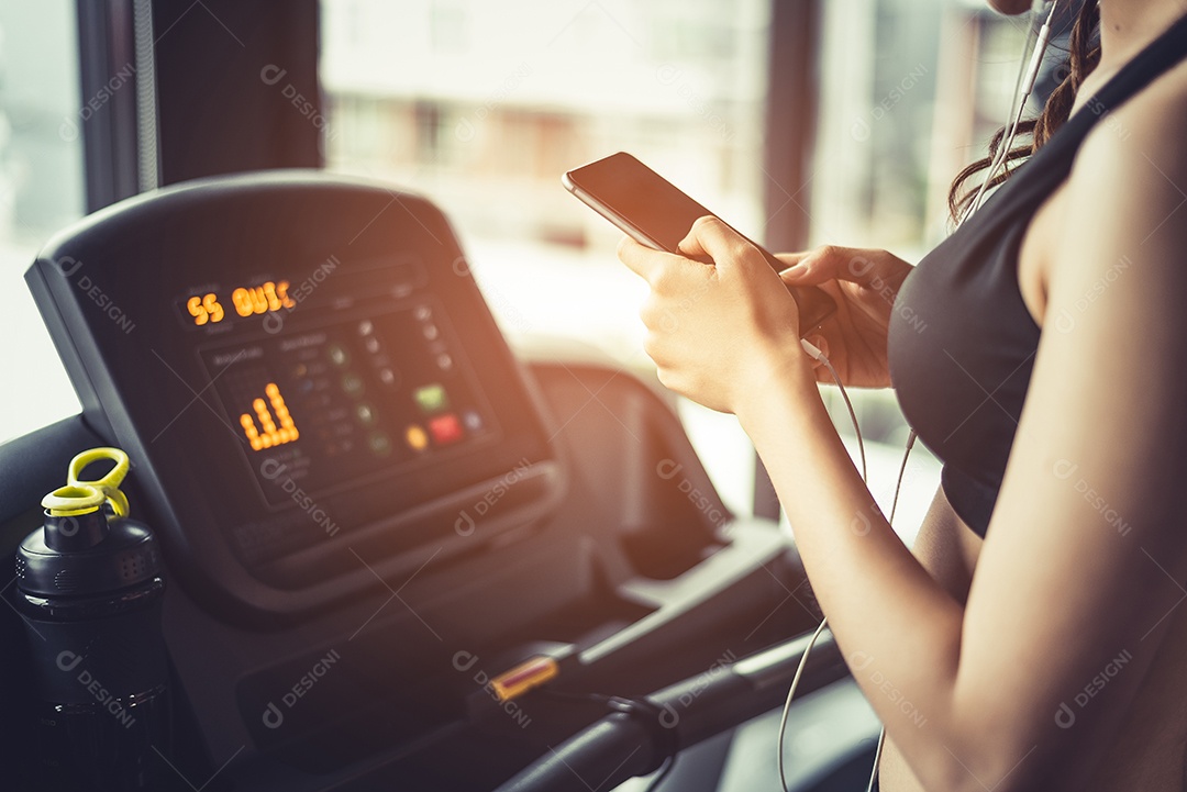 Mulher asiática usando telefone inteligente durante treino ou treinamento de força na academia de ginástica na esteira. Relaxe e o conceito de tecnologia.