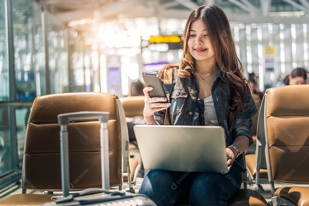 Passageira asiática jovem usando laptop e telefone inteligente enquanto está sentada no assento no saguão do terminal e esperando o voo no aeroporto.