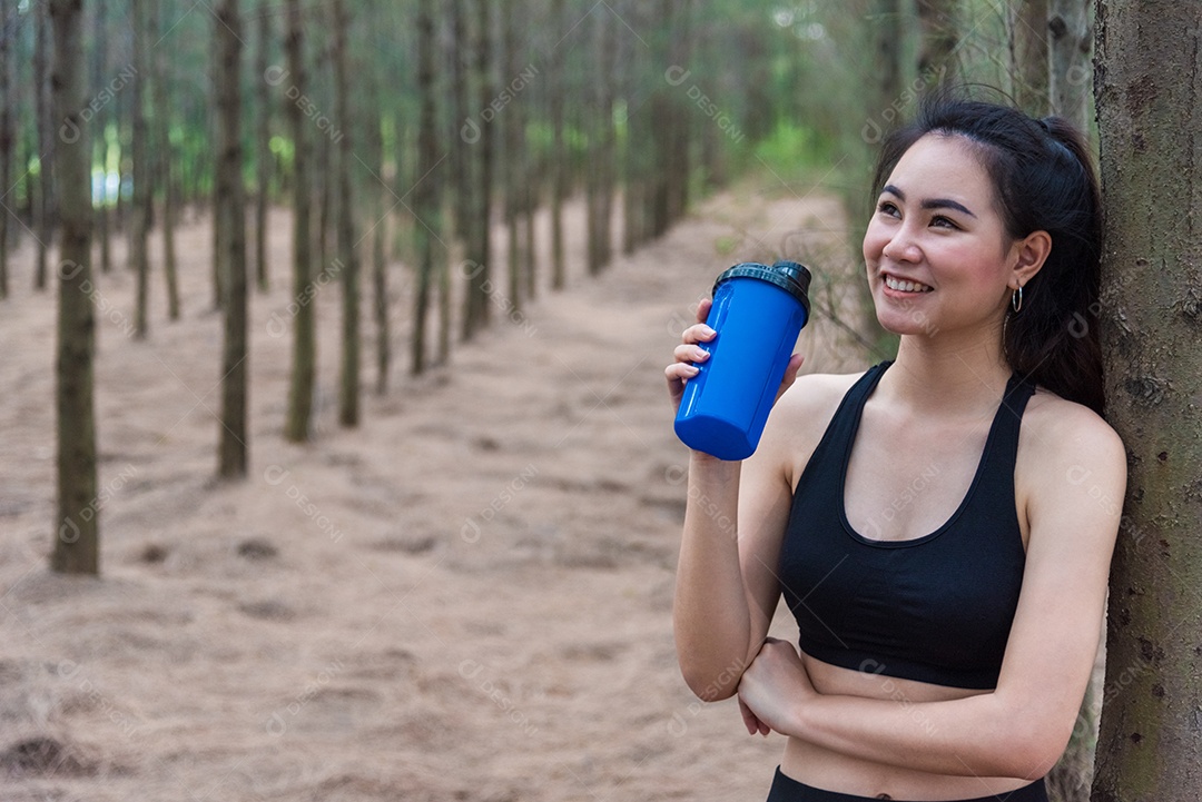 Mulher de esporte asiático de beleza descansando e segurando a garrafa de água potável e relaxando no meio da floresta depois de cansada de correr.