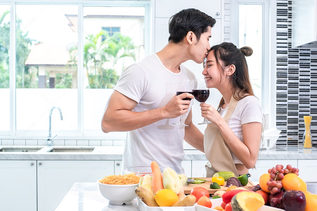 Amantes asiáticos ou casais beijando a testa e bebendo vinho na cozinha em casa.