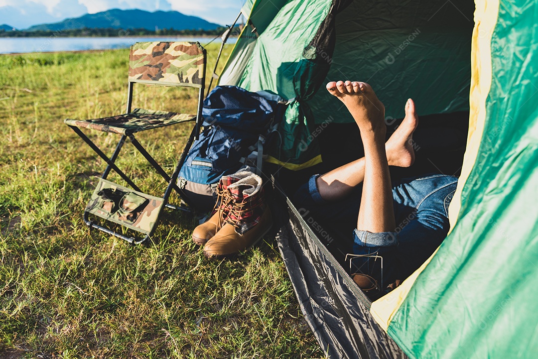 Feche as pernas da mulher relaxando na barraca de acampamento com lago de montanha e prado e fundo de campo de grama.