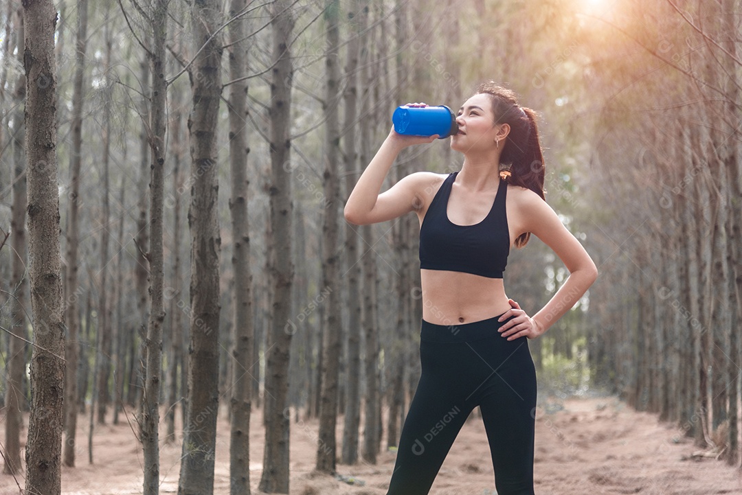 Mulher de esporte asiático de beleza descansando e segurando a garrafa de água potável e relaxando no meio da floresta depois de cansada de correr.