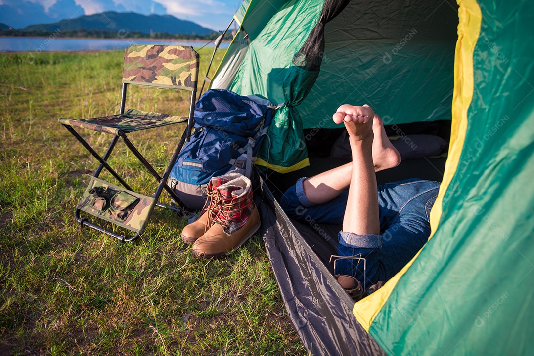 Feche as pernas da mulher relaxando na barraca de acampamento com lago de montanha e prado e fundo de campo de grama.