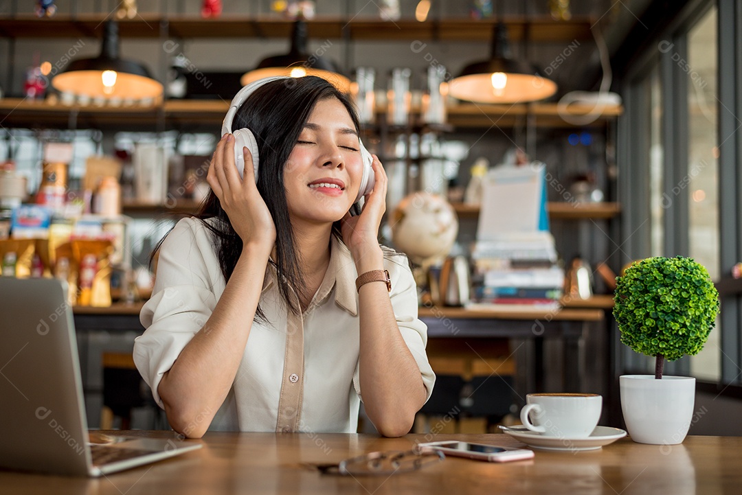 Mulher asiática feliz relaxando e ouvindo música na cafeteria com computador portátil e xícara de café.