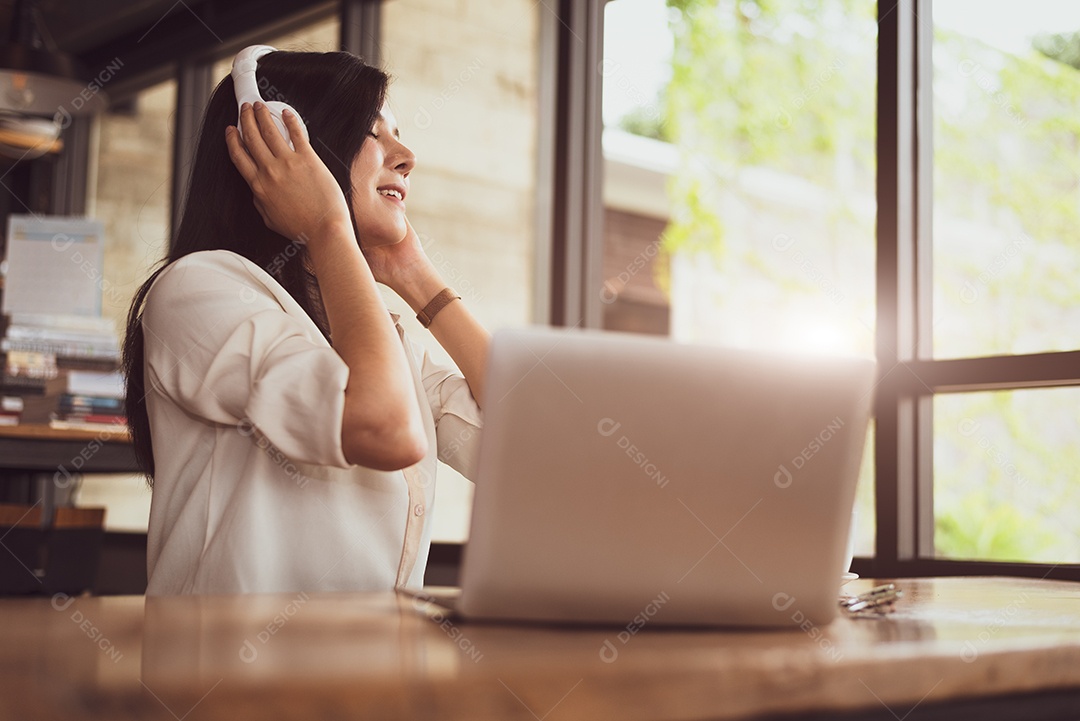 Mulher asiática feliz relaxando e ouvindo música na cafeteria com computador portátil.