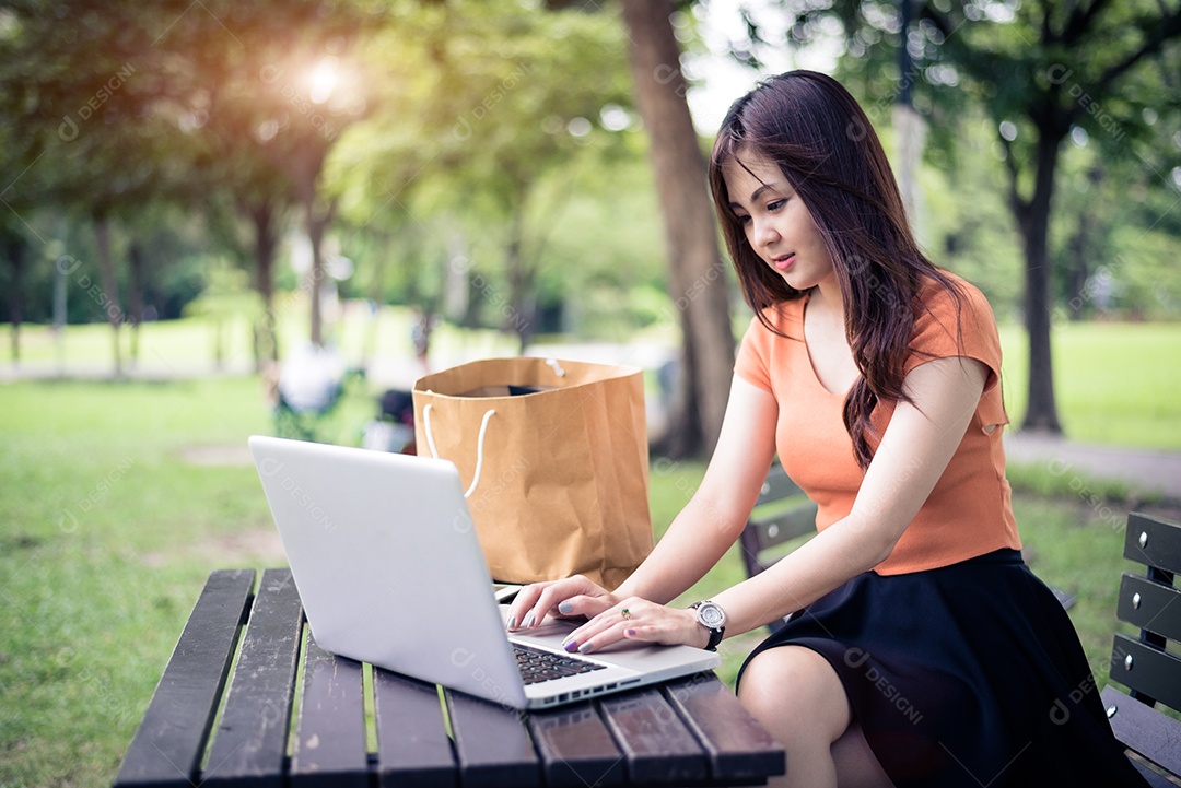 Mulher asiática usando e digitando no teclado do laptop no parque ao ar livre. Mulher conversando com seus amigos na rede social. Conceito de pessoas e estilo de vida. Tema de natureza e tecnologia.