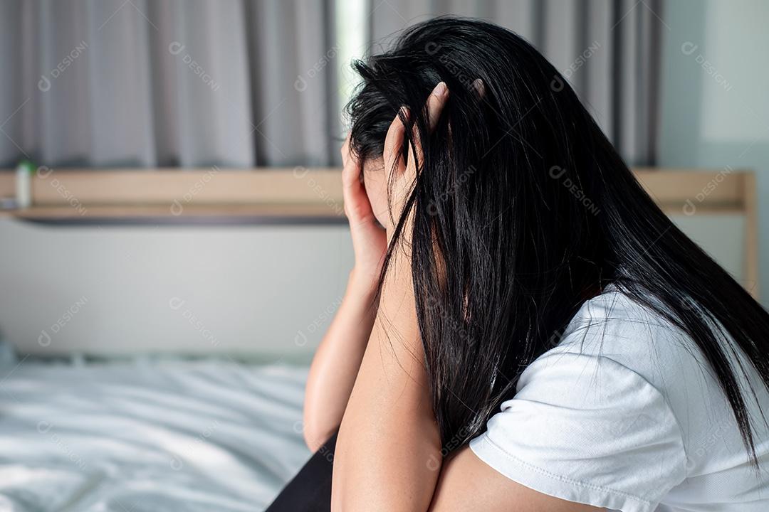 Mulher asiática de camisa branca, sentada na cama, segurando a cabeça e segurando um frasco de pílulas dietéticas para expressar estresse, decepção, depressão, problemas de saúde, cuidados de saúde e conceito de gerenciamento de estresse.