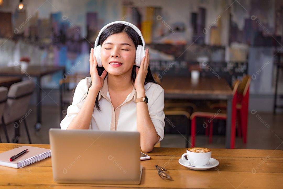 Linda mulher asiática relaxando e ouvindo música no café com computador portátil e xícara de café.