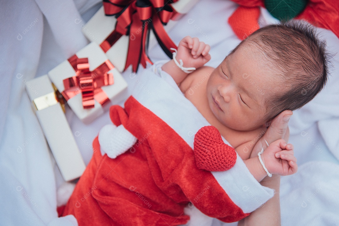 Bebê recém-nascido dormindo na mão da mãe no chapéu de Natal com caixa de presente do Papai Noel e coração de fio na toalha branca e macia.