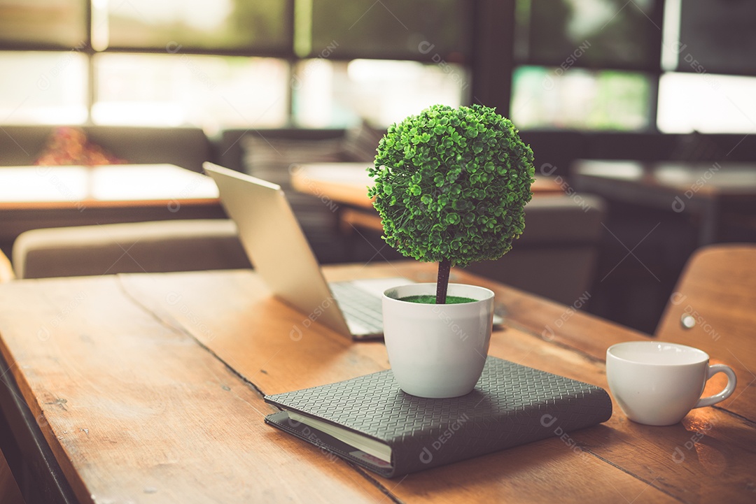 Árvore de decoração pequena, laptop, notebook e xícara de café na mesa de madeira na cafeteria.