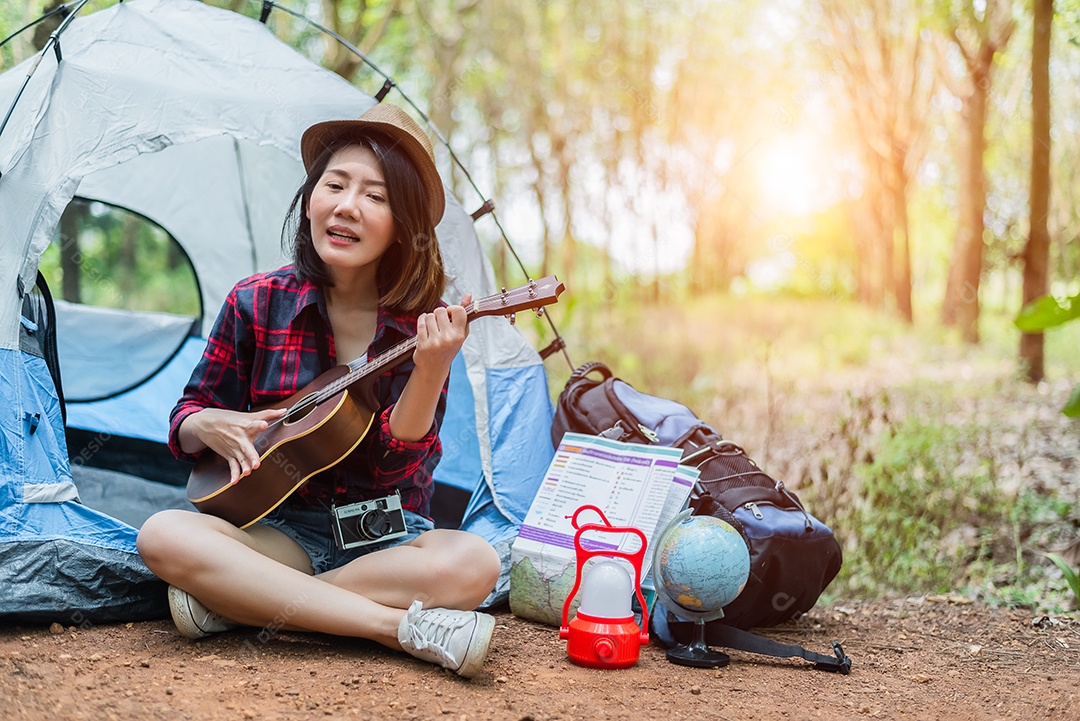 Linda mulher asiática tocando Ukulele na frente da barraca de acampamento.